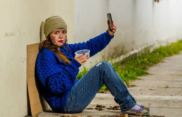 Vista al aire libre de la mujer sin hogar mendigando en la calle en el frío clima otoñal sentada en el suelo en la acera tomando una selfie —  Fotos de Stock