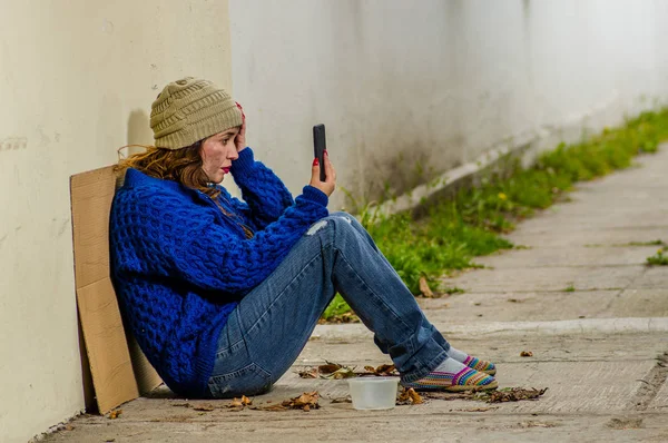 Vista al aire libre de una mujer sin hogar mendigando en la calle en el frío clima otoñal sentada en el suelo en la acera con un teléfono celular en la mano —  Fotos de Stock
