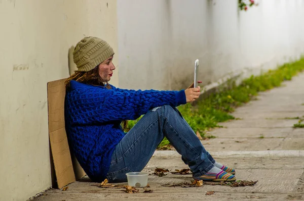 Outdoor view of homeless woman begging on the street in cold autumn weather sitting on the floor at sidewalk taking a selfie