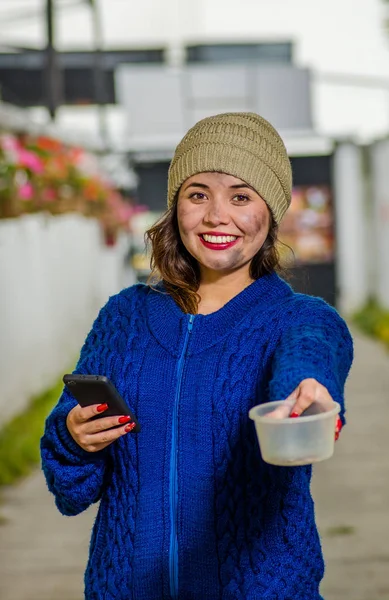 Zewnątrz widok bezdomne kobiety smutny na ulicy w zimnie jesień trzymając pustą kolbę z tworzywa sztucznego w jej ręce, z prośbą o pieniądze, przy użyciu telefon komórkowy na chodniku — Zdjęcie stockowe
