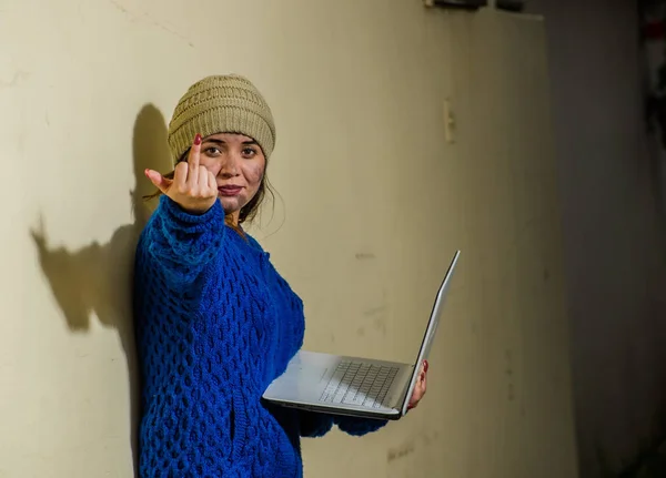 Vista ao ar livre da mulher sorridente sem-teto na rua em tempo frio outono segurando um computador em suas mãos e apontando ithe dedo médio na frente dela, na calçada — Fotografia de Stock