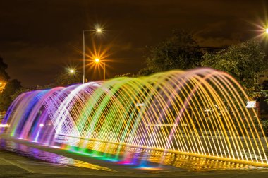 QUITO, ECUADOR- FEBRUARY 22, 2018: Beautiful outdoor view of colorful water entertainment structure fountain, at long exposure in the night, with buildings behind clipart