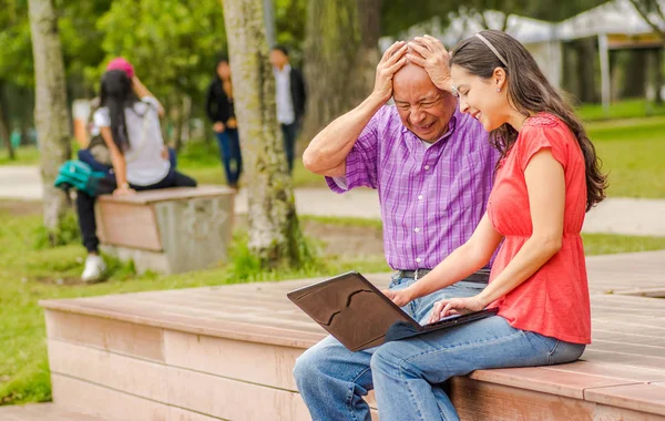 Outdoor view of father and daughter using a computer in the park, father do not understant how to use it