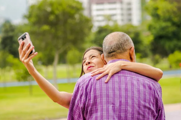 Zblízka dcery objímala svého otce, zatímco ona kontroluje její mobil na venku v parku — Stock fotografie