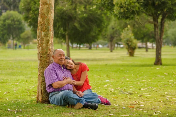 Вид на улицу, где дочь обнимает отца с любовью на открытом воздухе, сидя в траве, в парке — стоковое фото