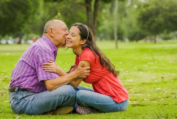 Vista ao ar livre do pai beijando sua filha com amor ao ar livre sentado no chão, no parque — Fotografia de Stock