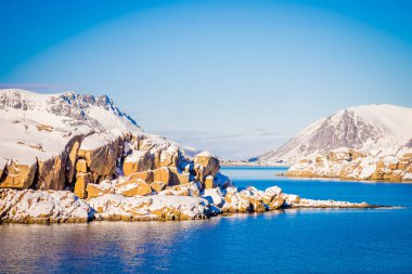 Outdoor view of snowy rock mountain covered during winter in the Arctic Circle, in a gorgeous blue sky and blue water clipart