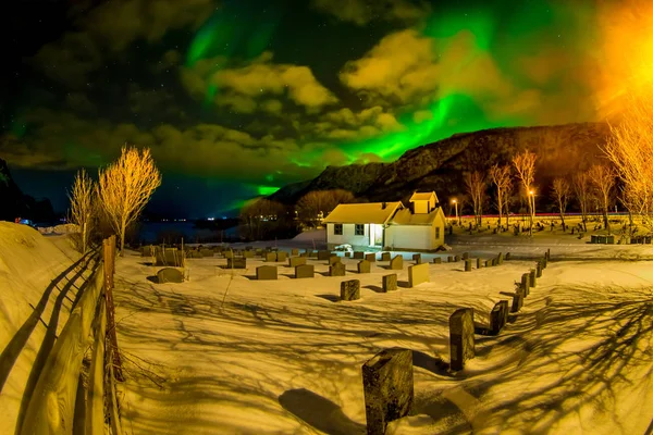 Vista al aire libre de la casa apedreada blanca con muchas lápidas cubiertas de nieve y hermosas luces del norte brillan brillantes y hermosas sobre picos afilados y lagos de las montañas Lofoten —  Fotos de Stock