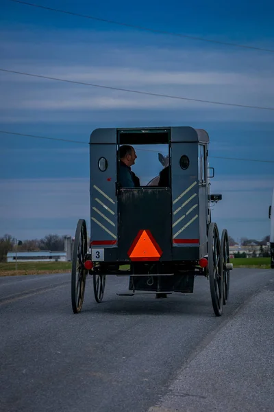 Pensilvania, Estados Unidos, 18 de abril de 2018: Vista al aire libre de la parte posterior de un buggy Amish anticuado con gente dentro y un caballo montando en un camino rural de grava — Foto de Stock