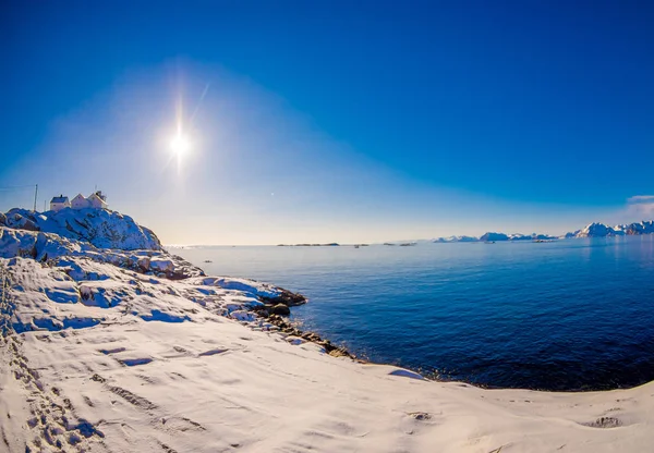 Vedere în aer liber a casei albe din lemn la țărm, toate terenurile acoperite cu zăpadă, soare strălucitor pe cer cu lac albastru în iarna înzăpezită în Cercul Arctic — Fotografie, imagine de stoc