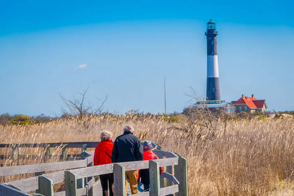 LONG ISLAND, EE.UU., 04 DE ABRIL DE 2018: Vista al aire libre de personas no identificadas caminando en un largo puente de madera en un hermoso día soleado con cielo azul con el faro de Montauk detrás, en Long Island — Foto de Stock