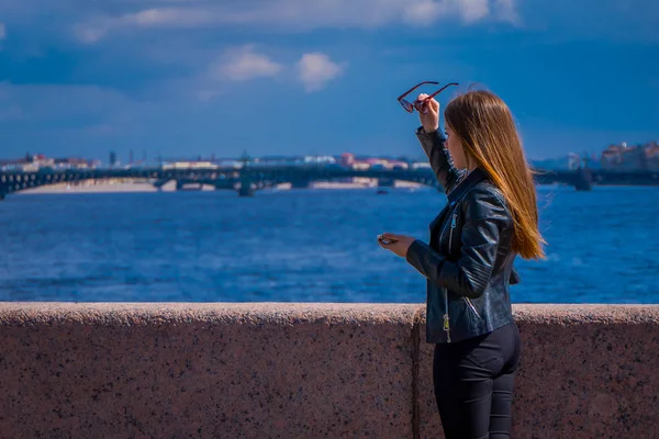 Kültéri nézet szőke orosz nő fekete ruhát visel, és a gazdaság egy napszemüveg azt a kezét a város St. Petersburg horizont homályos háttérrel — Stock Fotó