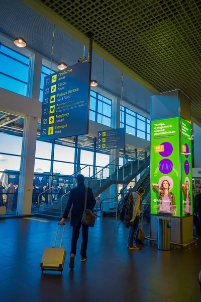 MINSK, BELARUS - 01 DE MAYO DE 2018: Vista interior de turistas no identificados caminando con sus maletas bajo un cartel informativo cerca de la zona de check-in dentro del aeropuerto de Minsk — Foto de Stock