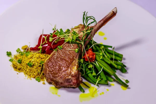 Placa de cerámica blanca con una chuleta de cordero, servida con judías verdes hervidas, cebolla coloreada con jugo de remolacha, pistacho amarillo de transporte, con brotes de romero y frijol sobre la superficie textil blanca. Vista lateral — Foto de Stock