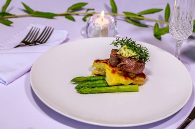 Angus bonfile Pommes Boulangerre, kuşkonmaz ve Paris tereyağı, biberiye, fasulye filizi ve genç pancar ile beyaz seramik tabak beyaz yüzey üzerinde bırakır. Yanan Mum arka plan üzerinde. Yan görünüm