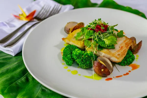 Placa de cerámica blanca con filete de Mahi-mahi y almejas con brócoli, cebolla coloreada con jugo de remolacha, perejil y brotes de frijol servidos sobre una hoja verde. Servilleta con dos tenedores y flor Strelitzia en el fondo . — Foto de Stock