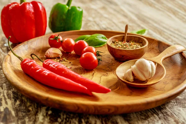 Fresh cherry tomatoes, garlic cloves, chili peppers, basil leaves, mix of dry spices in wooden pot with wooden spoon on wooden tray over wooden table, red and green capsicums on background. Side view — Stock Photo, Image