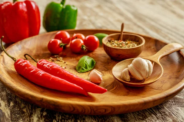 Fresh cherry tomatoes, garlic cloves, chili peppers, basil leaves, mix of dry spices in wooden pot with wooden spoon on wooden tray over wooden table, red and green capsicums on background. Side view