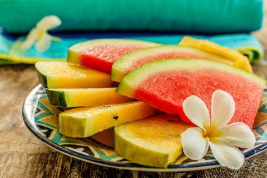 Kırmızı ve sarı karpuz Plumeria veya Frangipani çiçek renkli desenli seramik plaka üzerinde bir ahşap masa üzerinde dekore edilmiş dilimler kesin. Arka planda çizgili havuzu havlu. Yan görünüm