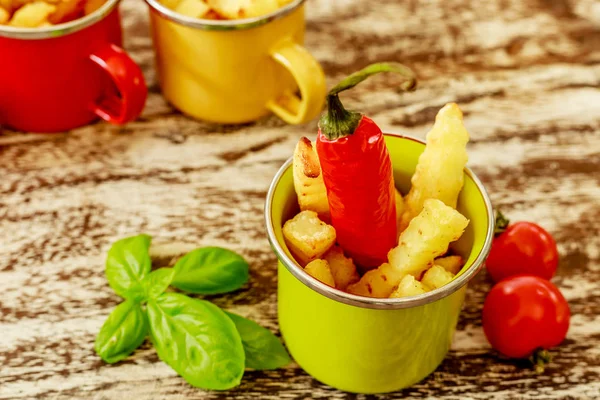 Green, yellow and red enameled cups with potato fries decorated with two cherry tomatoes, basil leaves and a red chilly pepper, over wooden table. Top view.