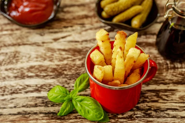 Red enameled cup with potato fries served with tomato sauce and pickled cucumbers in black ceramic pots and balsamic sauce in glass pot, decorated with basil leaves,over wooden table. Top view.