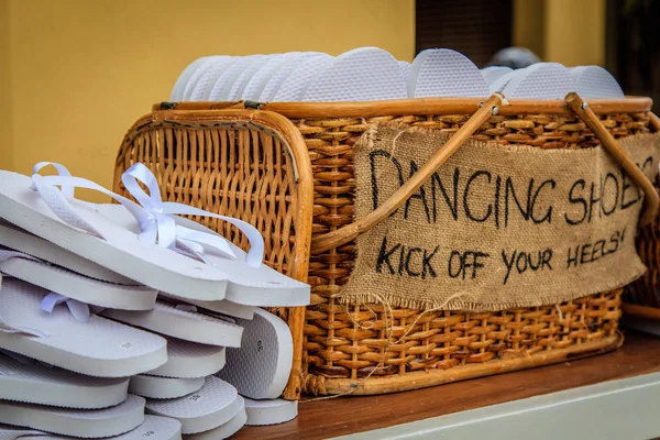 Rattan basket with white flip-flops of different sizes for guests, with a writing DANCING SHOES. KICK OFF YOUR HEELS!
