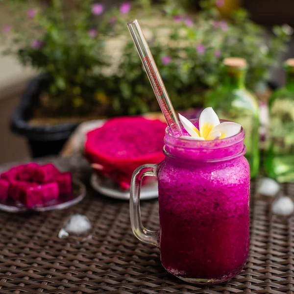 Red Dragon fruit juice in a glass with a glass straw decorated with Plumeria flower, cut dragon fruit on glass plates over dark brown fabric surface.