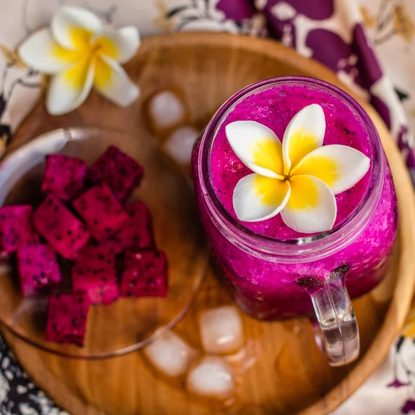 Red Dragon fruktjuice i ett glas, dekorerad med Plumeria blomma, skär dragon frukt och isbitar. Serveras på en runda träbricka över blommigt tyg. Ovanifrån. Kvadratisk bild. — Stockfoto