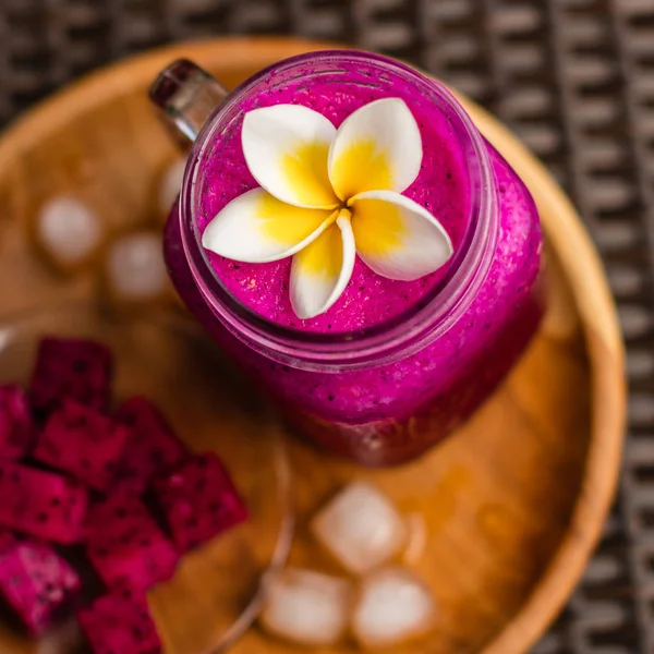 Red Dragon fruit juice in a glass, decorated with Plumeria flower, cut dragon fruit and ice cubes. Served on a round wooden tray over flowery fabric. Top view. Square image.