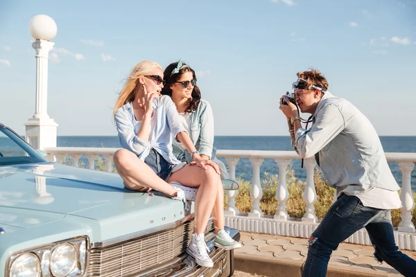 Man photographer taking photos of two women sitting on car — Stock Photo, Image