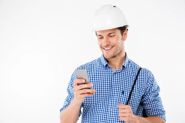 Sorridente jovem construtor em chapéu duro usando telefone celular — Fotografia de Stock