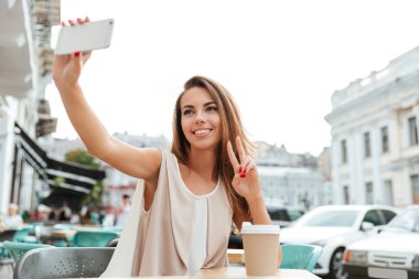 Güzel kız yapma selfie ve zafer gösterilen kafede oturum açın