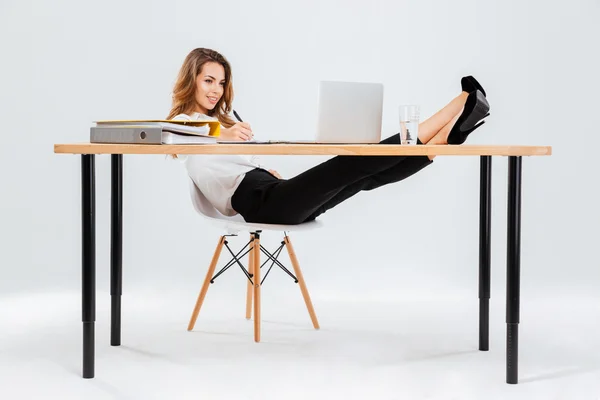 Mujer de negocios sonriente utilizando el ordenador portátil y la escritura con las piernas en la mesa — Foto de Stock