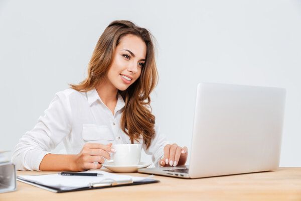 Happy attractive young businesswoman drinking coffee and using laptop