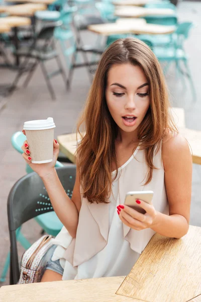 Surprised beautiful woman holding coffee cup and looking at smartphone — Stock Photo, Image