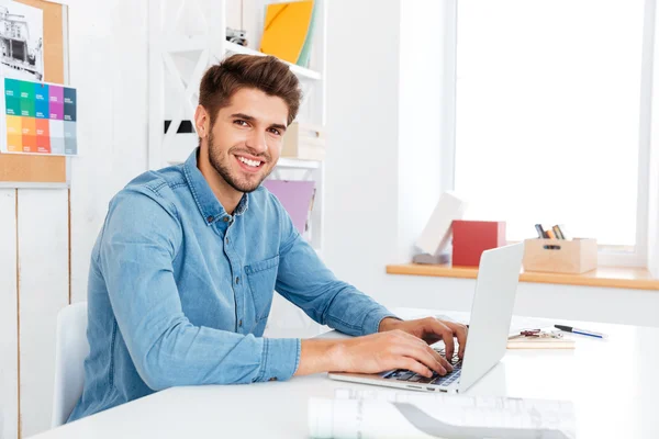 Joven hombre de negocios guapo escribiendo en el ordenador portátil y mirando a la cámara —  Fotos de Stock