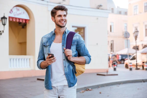 Hombre sonriente con mochila usando el teléfono celular en la calle — Foto de Stock