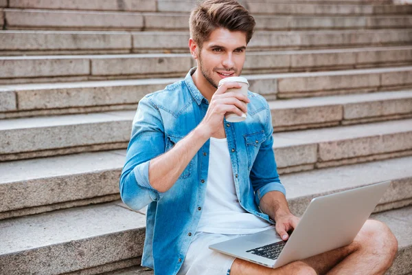 Hombre beber tomar tomar café y la celebración de la computadora portátil mientras está sentado — Foto de Stock