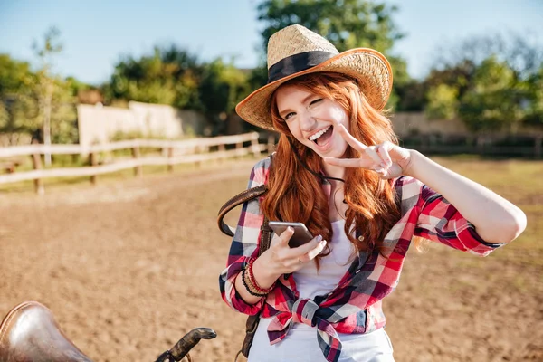 Vrolijke vrouw cowgirl met behulp van de mobiele telefoon en het vredesteken weergegeven: — Stockfoto