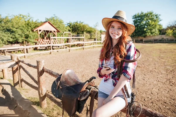 Улыбающаяся женщина ковбойша стоит и пользуется мобильным телефоном на ранчо — стоковое фото