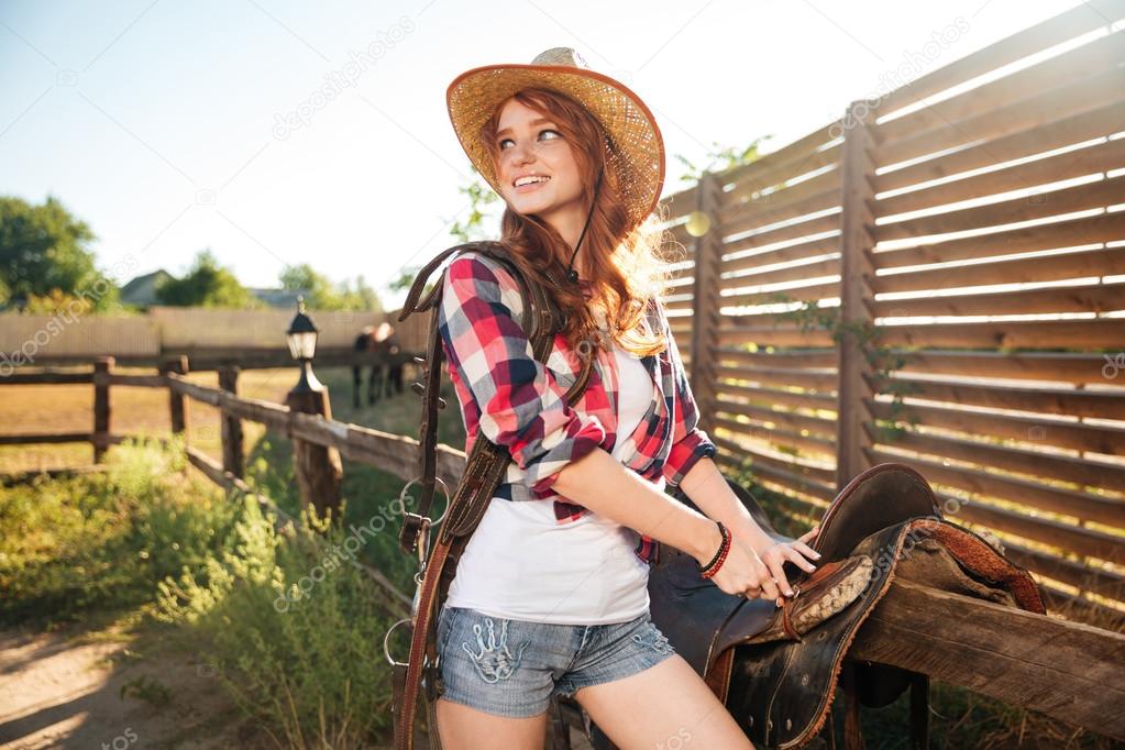 Happy redhead woman cowgirl preparing saddle for riding horse