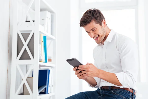 Riendo joven empresario sentado en la mesa y usando la tableta — Foto de Stock