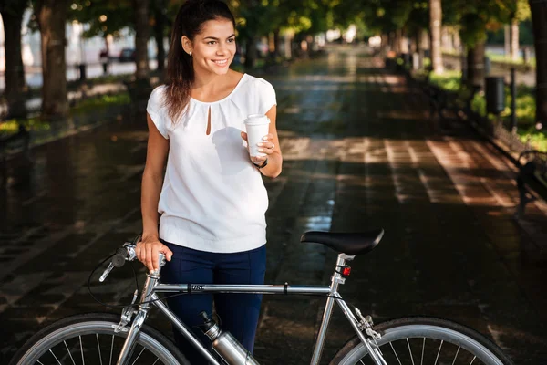 Женщина с чашкой кофе на вынос и велосипедом в парке — стоковое фото