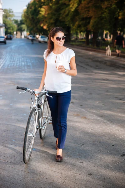 Mujer atractiva en gafas de sol caminando con bicicleta y café para llevar —  Fotos de Stock