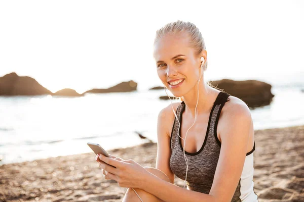 Senyum wanita olahraga pirang dengan earphone duduk di pantai — Stok Foto