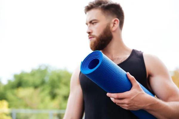 Serious male yoga instructor holding mat and looking away outdoors — Stock Photo, Image