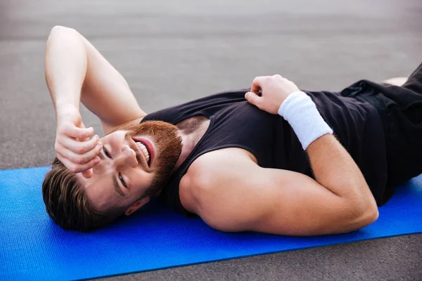 Alegre rindo esportes homem descansando no tapete de fitness azul ao ar livre — Fotografia de Stock