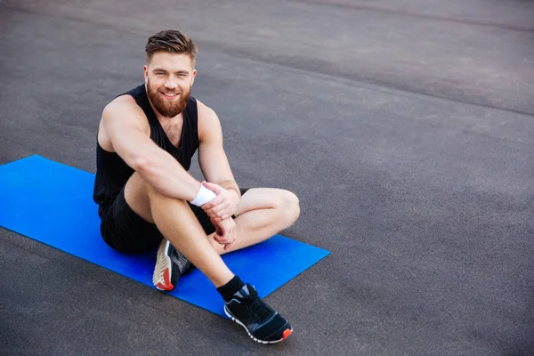 Homem de esportes sentado e descansando no tapete de fitness azul ao ar livre — Fotografia de Stock