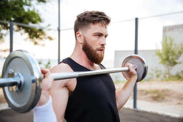 Serious young man athlete standing and holding barbell — Stock Photo, Image