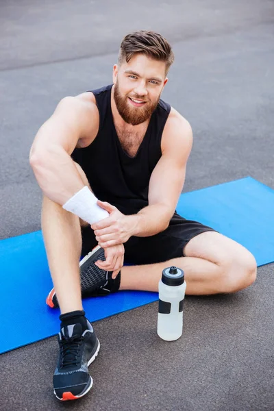 Sonriente joven deportista con botella de agua sentado y relajante — Foto de Stock